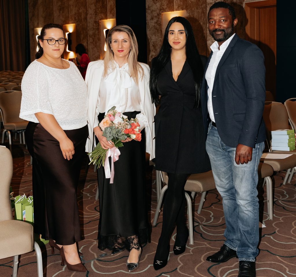 Four adults posing for a group photo at an indoor event; the woman in white holds a bouquet of flowers.