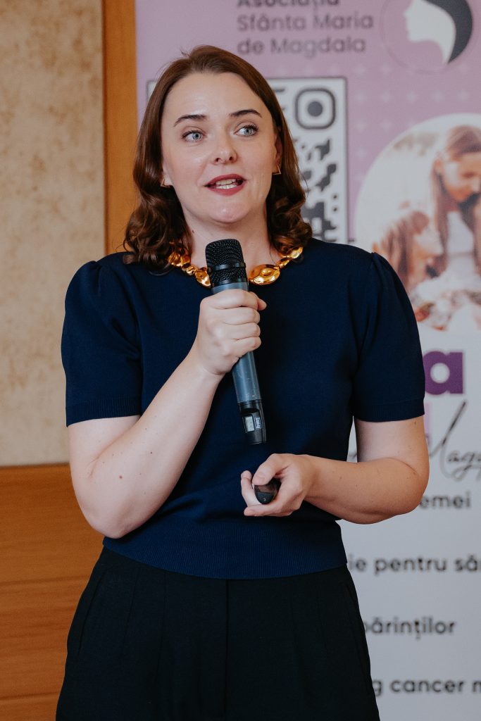 Woman in a navy dress speaks into a handheld microphone at a public event with a pink banner backdrop.