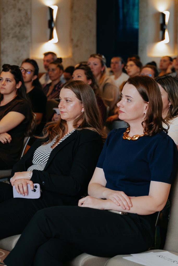 Two women in the front row of an indoor event, one holding a smartphone, with a focused audience behind them.