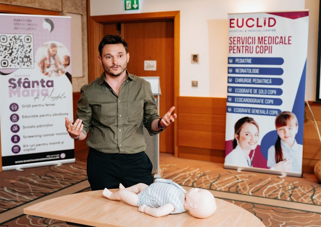 Man wearing green shirt explains CPR training with infant mannequins on a table, banners for medical services in the background.