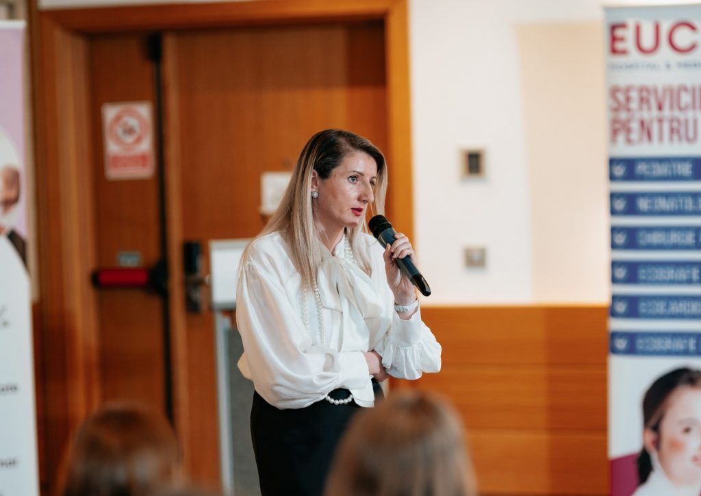 Woman in a white blouse and pearl necklace speaks into a handheld microphone at a conference, with an audience in the foreground.