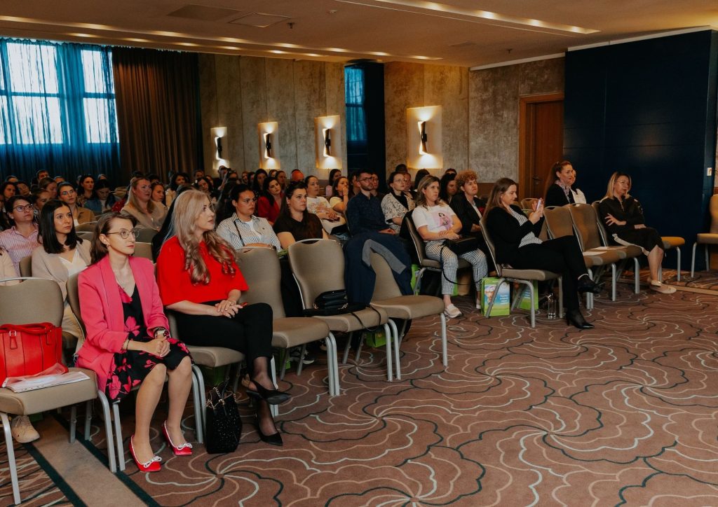 Audience of mostly women seated in rows of chairs watching a presentation in a conference room with patterned carpet and warm wall lighting.