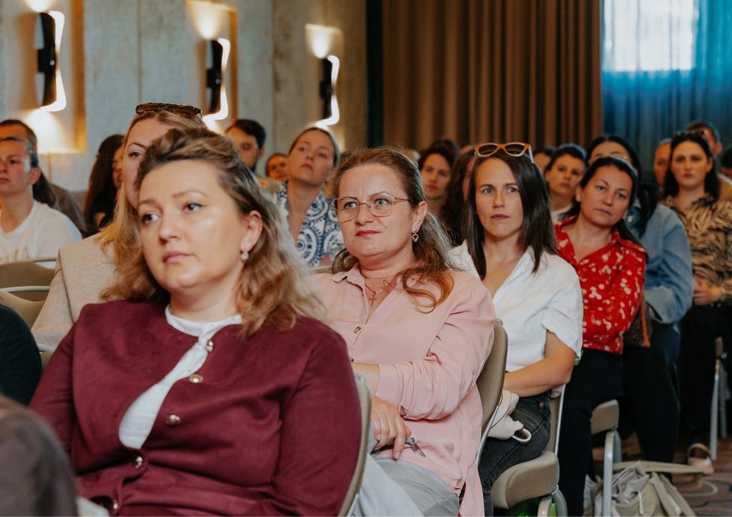 Audience of adults seated in rows at a conference or lecture, listening attentively in a dimly lit room
