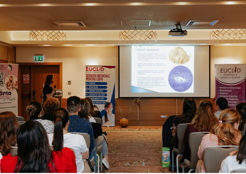 Speaker presenting a slide deck to an audience in a conference room with banners on stage.