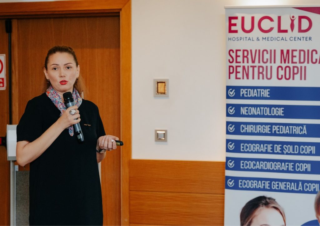 Woman speaking into a handheld microphone next to a tall banner for EUCLID Hospital & Medical Center with pediatric services listed
