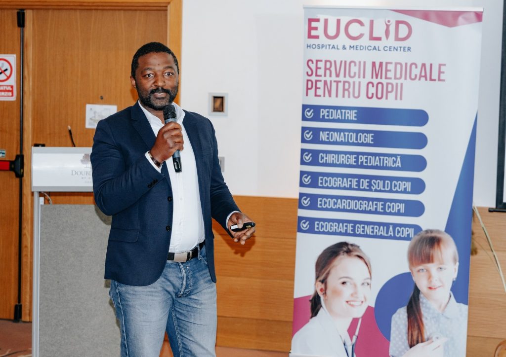 Male presenter in a navy blazer speaks into a handheld microphone beside a tall banner reading 'EUCLID Hospital & Medical Center' and listing pediatric services in Romanian.