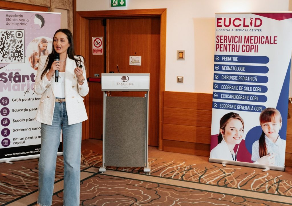 Female presenter speaks into a handheld microphone beside banners in a conference room.