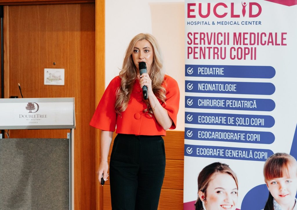 Woman in red blouse speaks into a microphone at a conference, with a tall banner listing pediatric medical services beside her.