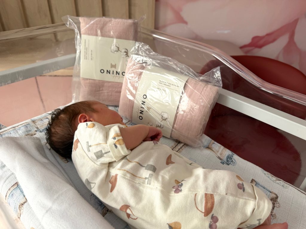 Newborn baby sleeping on a patterned blanket, wearing a light-colored onesie with animal prints, with a pink blanket and a clear plastic bag in the background.
