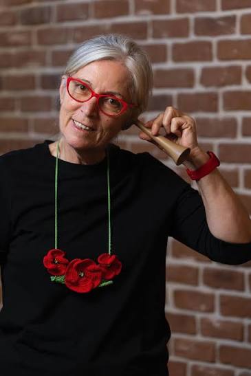 Senior woman with red glasses and a red flower necklace, standing against a brick wall, pretending to talk on a wooden rolling pin held to her ear.
